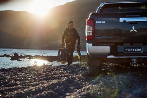 A Scuba diver walking on the rocks with his gear in hand towards his Triton ute
